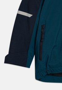 Jacket v odstínech modro-zelené a námořnické modři s kapsou na zip, texturovanou tkaninou a reflexním pruhem na rukávu, s nařaseným lemem.