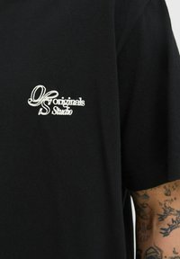 Camiseta de algodón negra con mangas cortas, con texto bordado en blanco: "DS Originals Studio," ubicado en la parte izquierda del pecho.