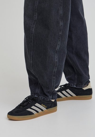 Melni zamšas Adidas Gazelle sporta apavi ar gumijas zolēm, ar baltām svītrām un zelta logotipu, saskaņoti ar tumšiem džinsa biksēm.