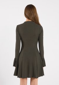 Kvinna med långt brunt hår som bär en åtsittande långärmad klänning i olivgrön färg med utsvängda ärmar, sedd bakifrån mot en vit bakgrund.