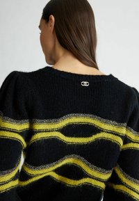 Femme aux longs cheveux bruns vue de dos portant un pull en maille noir avec des rayures horizontales jaunes et argentées métalliques.
