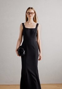 Black fitted dress with a square neckline and wide straps, textured fabric, falls to the floor. Model holds a small black bag.