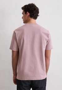 Młody mężczyzna z krótkimi kręconymi włosami, ubrany w gładką, jasnoróżową koszulkę z krótkim rękawem i ciemne spodnie, stojący przodem do białej, teksturowanej ściany.