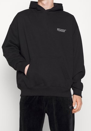 Person, die einen schwarzen Hoodie mit dem Schriftzug „PEGADOR“ auf der Brust und eine schwarze Hose trägt, eine Hand in der Kapuzen-Tasche, gezeigt vom Hals bis zu den Oberschenkeln.