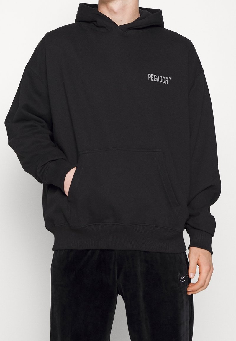 Personne portant un sweat à capuche noir avec le texte « PEGADOR » sur la poitrine et un pantalon noir, une main dans la poche du sweat, visible du cou aux cuisses.