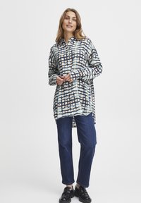 Camisa ligera de manga larga, tipo botón, en un patrón de cuadrícula azul, blanco y naranja; combinada con jeans oscuros y zapatos negros.