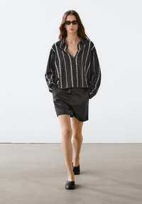 Zwart-wit gestreepte blouse met kraag en lange mouwen, gecombineerd met een zwarte rok en zwarte ballerina's, op een neutrale achtergrond.