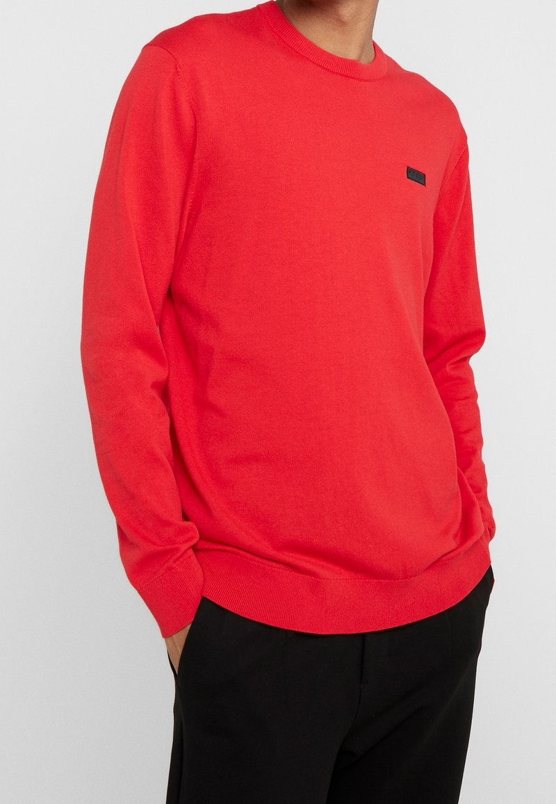 Homme portant un pull rouge vif à manches longues et un pantalon noir, mains dans les poches sur un fond neutre.