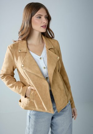 Kobieta o brązowych włosach, ubrana w beżową zamszową kurtkę motocyklową na białej koszuli zapinanej na guziki i jasnoniebieskich dżinsach, pozująca na szarym tle.