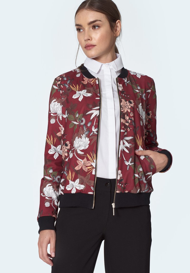 Kwiatowa kurtka bombowca na maroonowym tle, z czarnymi ściągaczami przy mankietach i u dołu, złotymi zamkami oraz kontrastującymi białymi kwiatami i liśćmi.