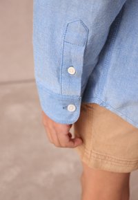 Vaaleansininen denimpaita, jossa on napillinen kalvosin ja olkasauman yksityiskohta, yhdistetty ruskeisiin shortseihin, korostaen kankaan rakennetta ja ompeleita.