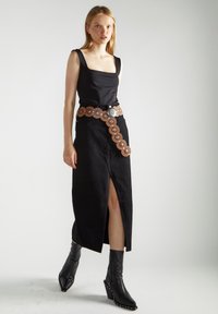 Abito nero senza maniche con scollo a quadrato e uno spacco laterale, abbinato a una cintura decorativa marrone con accenti circolari e una fibbia argentata. Stivaletti neri con tacco a zeppa completano il look.
