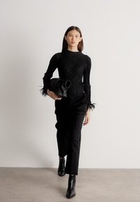 Sieviete staigā melnā ribotā topā ar spalvu manžetēm, melnās biksēs, ar sānu punktveida zābakiem, turot rokās lielu melnu aploksnes tipa somiņu.