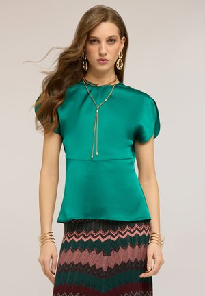 Kobieta o długich falowanych włosach, ubrana w satynową bluzkę w kolorze morskim, spódnicę w zygzakowe wzory, złote kolczyki koła, warstwowe naszyjniki oraz liczne złote bransoletki.