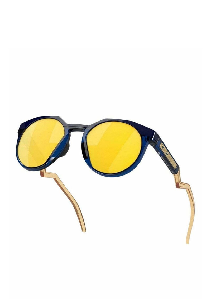 Lunettes de soleil bleu et or avec un design géométrique. Elles sont dotées de verres jaunes et de branches angulaires uniques, combinant des matériaux en plastique et en métal.