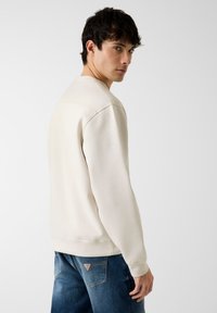 Sweat-shirt beige clair à col rond et manches raglan, associé à un jean bleu foncé présentant un patch logo sur la poche arrière.