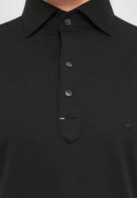 Svart polo skjorte med krage, tre svarte knapper og liten brodert "MK"-logo på brystet.