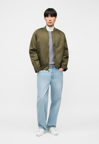 Νέος άνδρας στέκεται με τα χέρια στις τσέπες, φοράει μπουφάν bomber σε ελαιί πράσινο, γκρι πουλόβερ, ανοιχτού μπλε τζιν και λευκά αθλητικά παπούτσια σε λευκό φόντο.