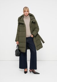 Olive green puffer coat with a hood, featuring a belted waist, paired with a beige turtleneck and wide-leg dark denim jeans. Black handbag and heels.