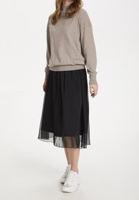 Beige stickad tröja med hög halsringning, matchad med en svart chiffong-midi kjol. Vita sportskor fulländar outfiten.