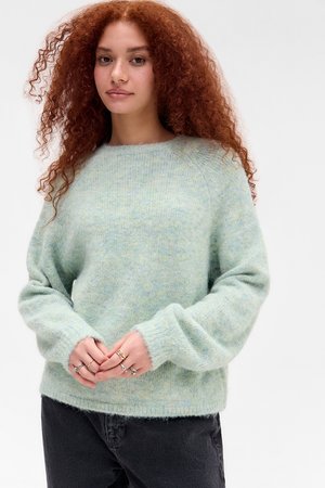 Jong iemand met lang, krullend, rood haar, gekleed in een zachte lichtblauwe trui en zwarte broek, met de handen gevouwen en meerdere ringen zichtbaar.