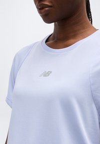 Γυναίκα που φοράει αθλητική μπλούζα κοντομάνικη σε ανοιχτό μωβ χρώμα με αντανακλαστικό λογότυπο New Balance στο κέντρο του στήθους.