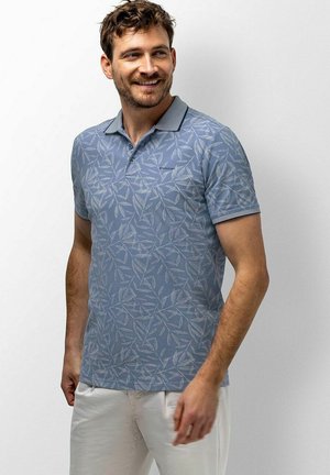 Lächelnder Mann, der ein blaues kurzärmeliges Poloshirt mit Blattmuster und weiße Hose trägt, steht vor einem einfarbigen hellen Hintergrund.