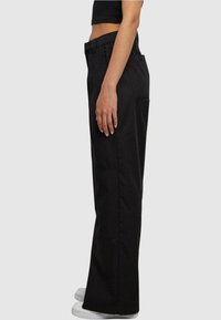 Personne portant un pantalon noir taille haute à jambes larges et des baskets blanches, debout de profil sur un fond clair uni.