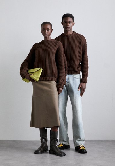 Suéteres de punto acanalado marrones, una falda midi caqui, botas de cuero negras y un bolso de mano verde vibrante. La modelo está al lado de una figura con jeans claros y zapatos.