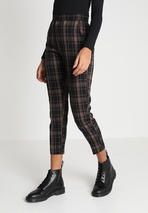 Nainen, jolla on mustat ja ruskeat ruudulliset nilkkapituiset housut sekä mustat nauhalliset nilkkurit, seisomassa yksinkertaisen vaalean taustan edessä.