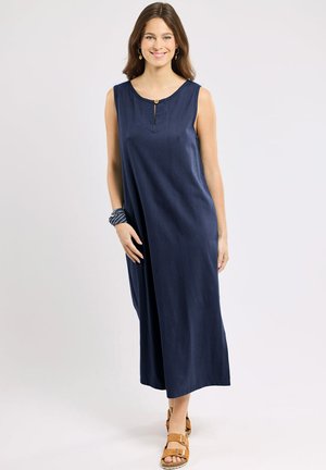 Femme debout et souriante portant une robe midi bleu marine sans manches avec un col boutonné et des sandales beige.