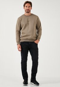 Sudadera beige con cuello redondo, puños y dobladillo acanalados, con un sutil logo negro en el pecho, combinada con vaqueros azul oscuro y zapatillas negras.