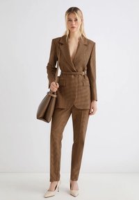 Blazer a scacchi marrone con vita cintata, abbinato a pantaloni coordinati. Tacchi chiari e una borsa rigida marrone completano il look.
