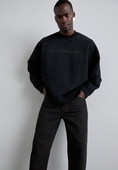 Must mustriga särkk, millel on kulumisjäljed ja "WON HUNDRED" tekst. Kombineeritud tumedate teksadega, luues monokroomse üldilme.