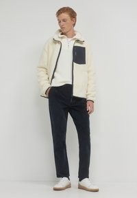 Giovane uomo con giacca in pile crema con tasca nera, pullover bianco texturizzato, pantaloni neri e scarpe da ginnastica bianche, in piedi contro uno sfondo bianco.