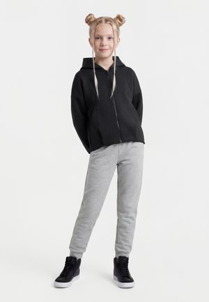 Junges Mädchen mit blondem Haar, das zu zwei Dutts und Zöpfen frisiert ist, trägt einen schwarzen Kapuzenpullover mit Reißverschluss, graue Jogginghosen und schwarze Turnschuhe und steht mit hinter dem Rücken verschränkten Händen.