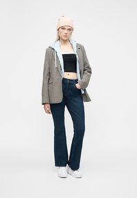 Νέα γυναίκα με μπεζ σκουφί, γκρι καρό σακάκι, ανοιχτογάλαζιο φούτερ με κουκούλα, μαύρο κοντό τοπ, μπλε τζιν και λευκά αθλητικά παπούτσια, στέκεται με το ένα χέρι στην τσέπη.