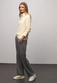 Krémový rolákový svetr z měkkého úpletu, v kombinaci se šedými širokými kalhotami s jemným proužkem a světlými teniskami, ruce v kapse, stojí před neutrálním pozadím.