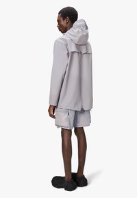Jasnoszara, wodoodporna kurtka z kapturem, o luźnym kroju i detalach wentylacyjnych z tyłu, zestawiona z jasnoszarymi szortami i fakturowanymi czarnymi butami.