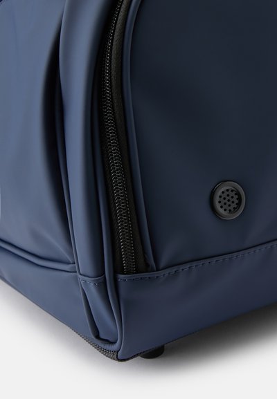 Mochila azul marinho com acabamento mate suave, fecho preto e um buraco de ventilação circular com textura de malha na lateral.