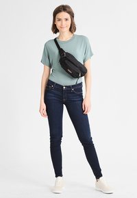 Musta vyölaukku vetoketjulla, joka on päällystetty vaaleanvihreän paidan ja tummansiniset farkut, yhdistettynä beigen värisiin tennareihin. Yksinkertainen muotoilu ja värit.