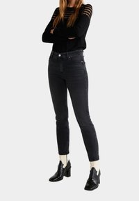 Jean skinny noir taille haute, associé à un haut noir à manches longues avec des découpes horizontales et des bottines noires brillantes.