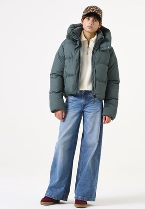Grüne Daunenjacke mit hohem Kragen, kombiniert mit einem cremefarbenen Pullover, blauen weiten Jeans und burgunderfarbenen Wildlederschuhen. Leopardmuster-Basecap.