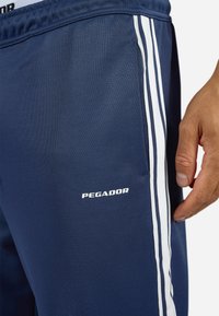 Marineblå træningsbukser med hvide striber, der har en sidelomme og "PEGADOR"-logo i hvid på venstre ben. Blødt, glat stof.