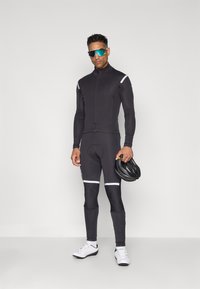 Tenue de cyclisme noire à manches longues en matériau extensible ; comporte des bandes réfléchissantes, une fermeture éclair à l'avant et des chaussures de cyclisme ; tenant un casque.