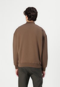Brown sweatshirt with a high collar, dropped shoulders, and ribbed cuffs, paired with dark green pants. Smooth, cotton-like texture.