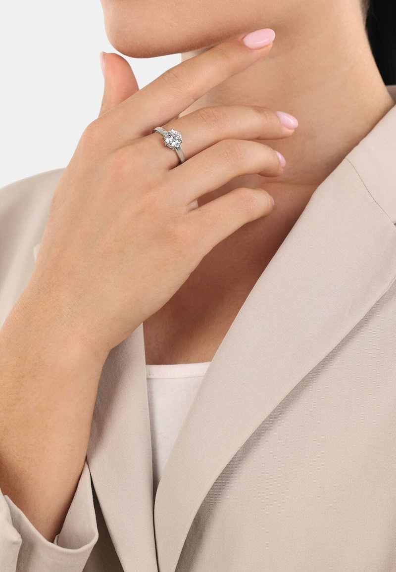 Silverring med en central klar ädelsten, bärs på en hand som rör vid hakan. Mjuk beige blazer och ljusrosa nagellack synligt.