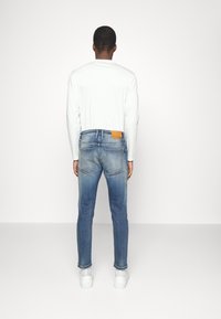 Blå denimjeans med en urtvättad effekt, med en beige läderlapp vid midjan och en slim fit-design. Bärs tillsammans med en vit långärmad skjorta.