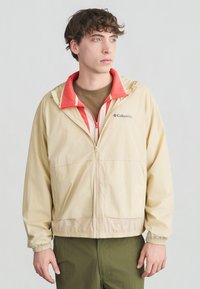 Νέος άνδρας που φοράει μπεζ μπουφάν Columbia με κόκκινη εσωτερική επένδυση και χακί παντελόνι, κοιτάζει προς τα πλάγια σε ένα απλό φόντο.