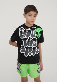 T-shirt nera in cotone con grafica aliena bianca e verde; abbinata a pantaloni corti neon verdi con stampe di cartoni grigi.